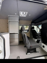Interior of a modern RV rental van featuring a rear bench seat, stainless fridge, ceiling fan, and compact kitchen cabinets.