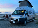 An RV rental camper van with a pop-top is parked by a lake at twilight, two children are at a picnic table nearby.