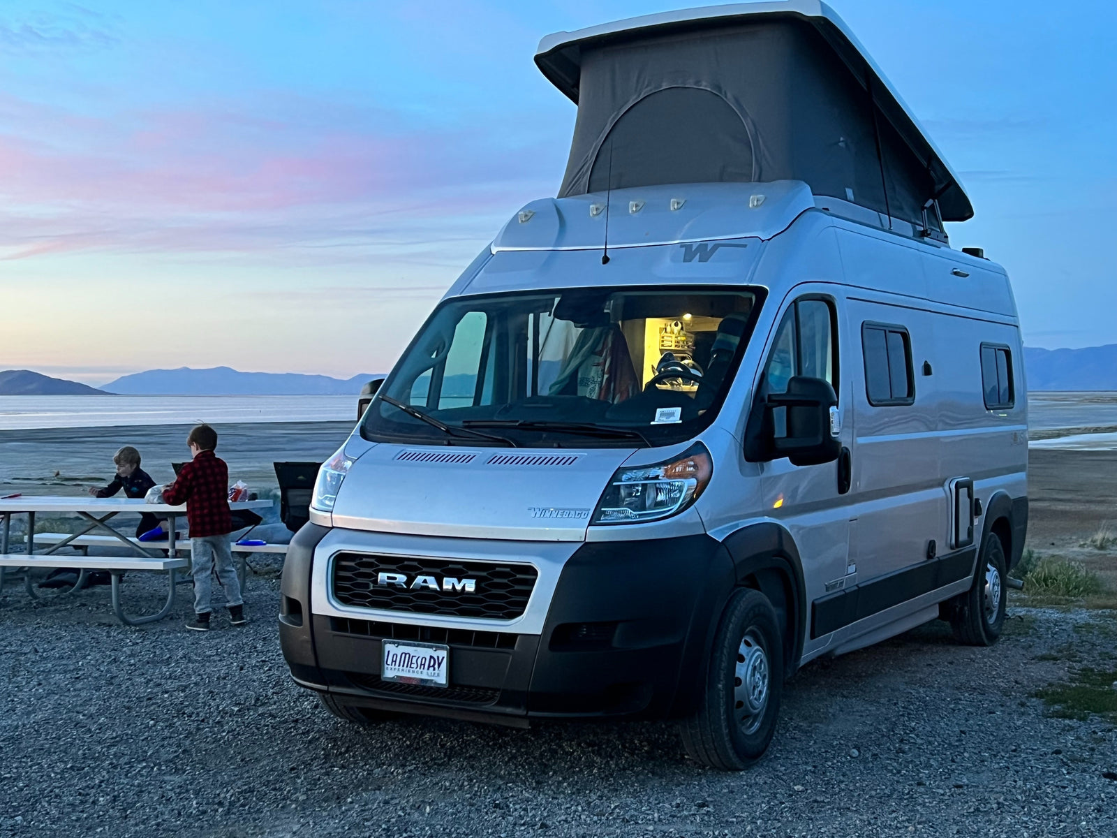 An RV rental camper van with a pop-top is parked by a lake at twilight, two children are at a picnic table nearby.