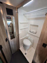 Compact RV rental bathroom featuring a toilet, shower, built-in shelves, and a mirror.
