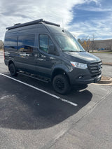 Dark gray Mercedes-Benz Sprinter camper van, ideal for RV rental adventures, parked in a lot under a cloudy sky.