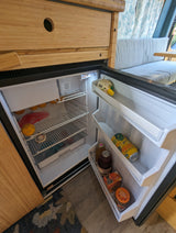 Open compact RV rental fridge with oranges, canned drinks, baking soda, and marmalade for your mobile journey.