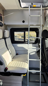 RV rental interior with white quilted seats, a scenic window view, overhead storage, and a ladder to an upper bunk.