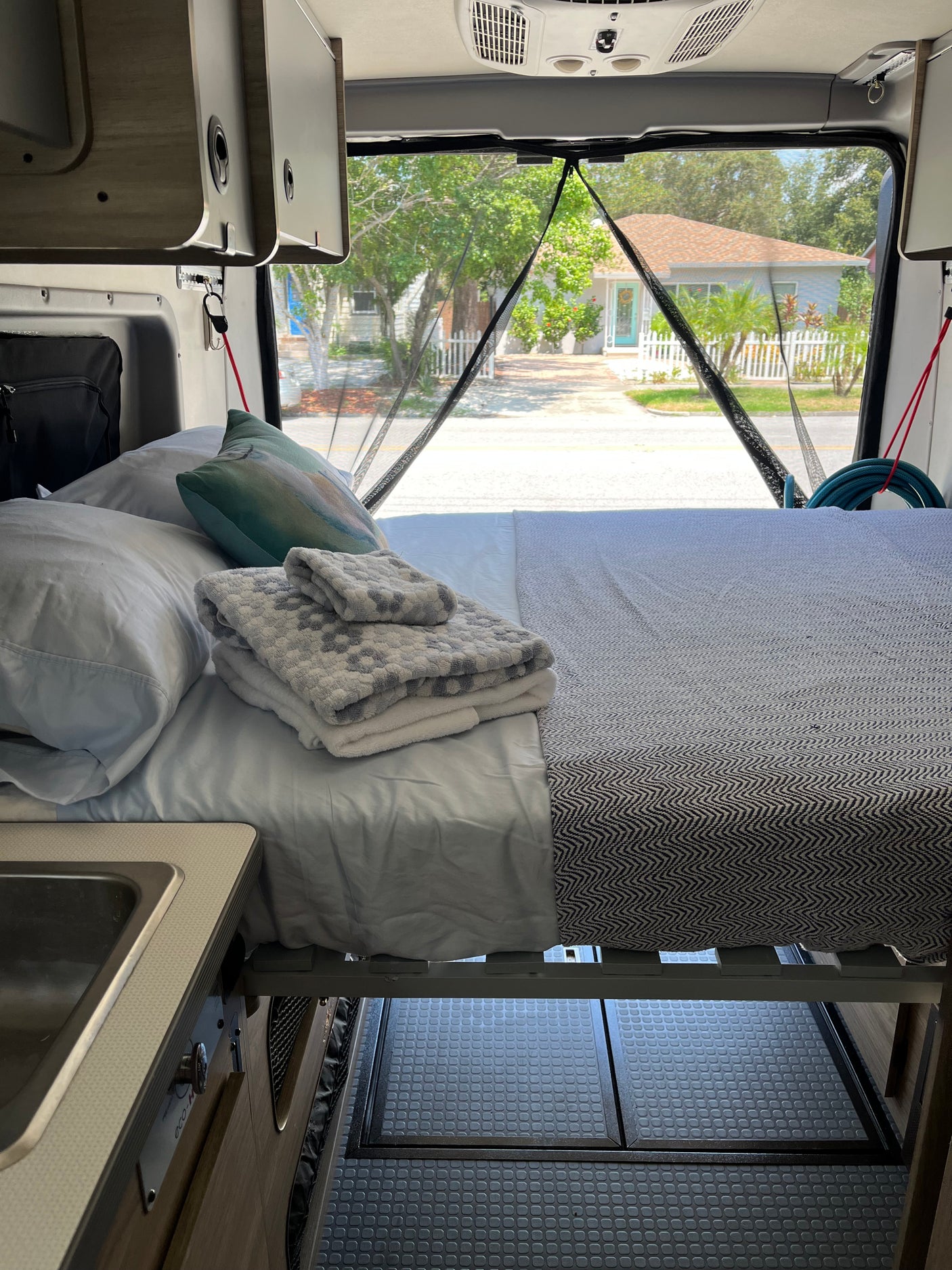 RV rental interior featuring a cozy bed, kitchenette, and a view of a charming street through the mesh screen.