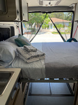 RV rental interior featuring a cozy bed, kitchenette, and a view of a charming street through the mesh screen.