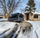 Gray RV rental van on a snowy driveway leading to a large house. Bare trees and winter landscape visible.