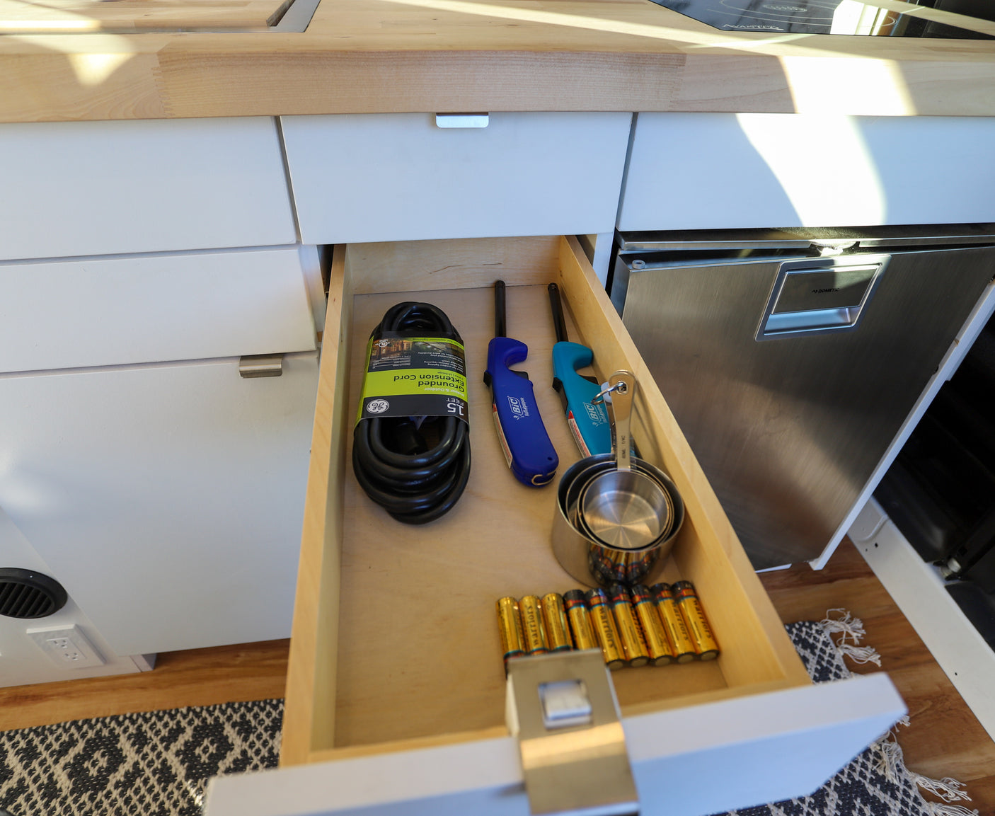 Compact RV rental kitchen with an open drawer revealing a GE extension cord, Bic lighters, measuring cups, and Dometic fridge.