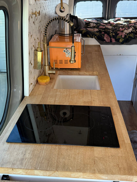 Inside an RV rental: a stylish compact kitchen with wood countertops, sink, induction cooktop, and gold fixtures for van life.