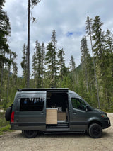 A sleek grey camper van, ideal for an RV rental, with its side door open revealing a light wood interior, parked in a dense forest.