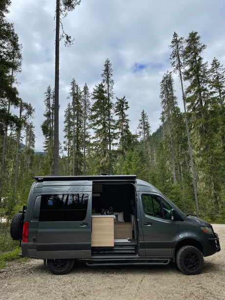 A sleek grey camper van, ideal for an RV rental, with its side door open revealing a light wood interior, parked in a dense forest.