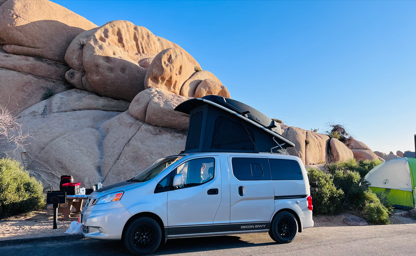 A silver pop-top RV rental camper van parked in a sunny, rocky desert campsite. A green tent is nearby.