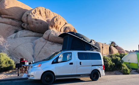 A silver pop-top RV rental camper van parked in a sunny, rocky desert campsite. A green tent is nearby.