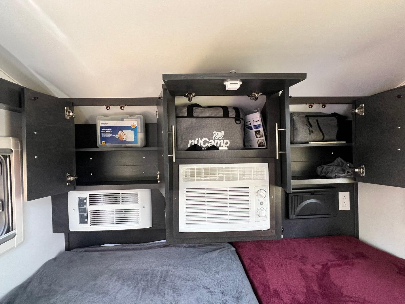 RV rental interior: organized overhead cabinets revealing a first aid kit & branded bag, dual AC units, and plush bedding for two.