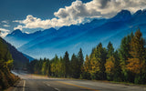Scenic RV rental journey on a mountain road, flanked by vibrant autumn forests and layered blue peaks under a cloudy sky.