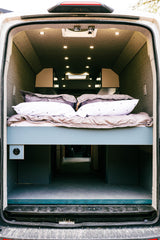 Rear view inside a modern RV rental, featuring a raised bed with pillows and sheets, and a large storage area below.