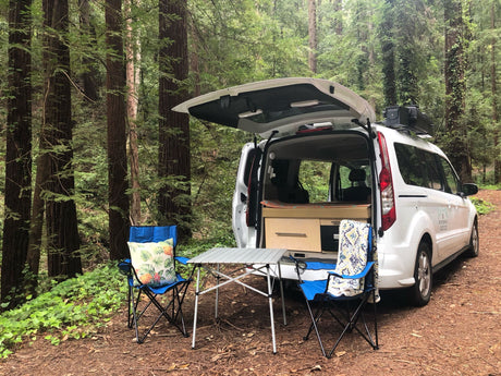 This RV rental shows a camper van in a redwood forest, with a comfortable outdoor setup: chairs & table for camping.