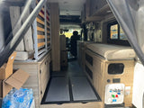 RV rental camper van interior: folded bed frame, stacked cushions, bench seating, and water utility hookups.