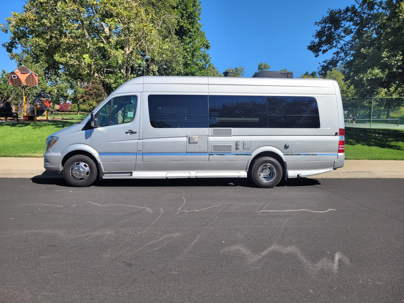  seen parked on a sunny street.|Silver RV rental parked on a sunny day. A park with a playground and tennis courts is visible nearby.|Comfortable cream captain's chairs inside an RV rental. Explore mobile living with this well-appointed camper van interior.|RV rental interior: Looking towards the back — photo 8