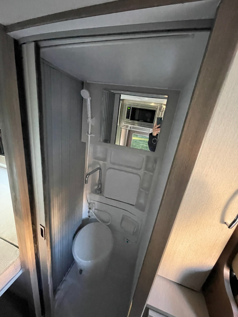 Inside this RV rental, view a compact wet bath with toilet, sink, and shower. A mirror reflects the kitchen area. — photo 5