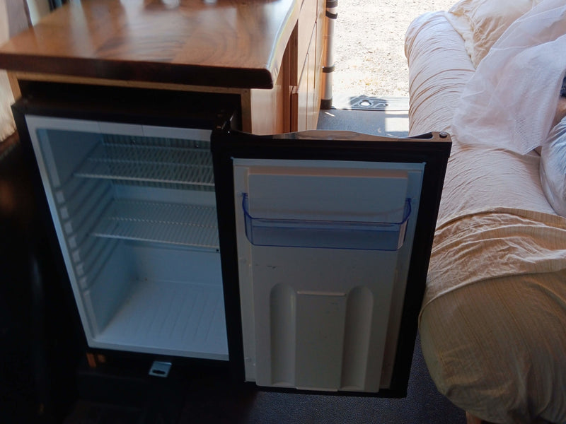 An open compact fridge, part of the functional interior of an RV rental, positioned below a wooden counter, next to a cozy sleeping area. — photo 7