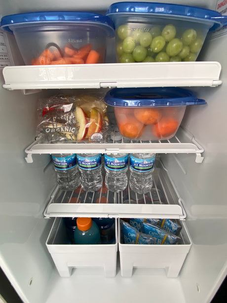 RV rental fridge stocked with carrots, grapes, apples, mandarins, and water bottles – healthy road trip snacks.
