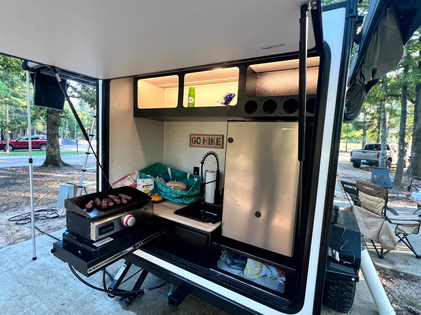 Outdoor kitchen of an RV rental, complete with a griddle cooking sausages, sink, fridge, and shelves for camping. — photo 5