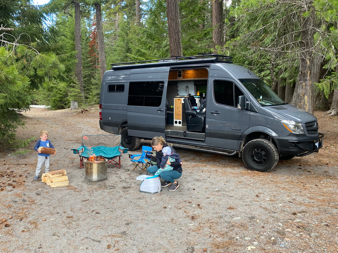 Family camping around a campfire with their Sprinter RV rental in a serene forest.