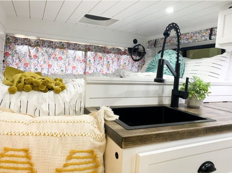 Inside a stylish RV rental: a modern kitchenette with a black sink, a cozy bed, and floral curtains.
