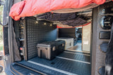 Spacious RV rental interior with overhead bed, storage net, control panel, and large gear box on textured floor.