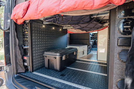 Spacious RV rental interior with overhead bed, storage net, control panel, and large gear box on textured floor.