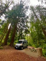 Explore Washington's nature! This RV rental campervan is set up at a serene forest campsite with a stone fire pit.