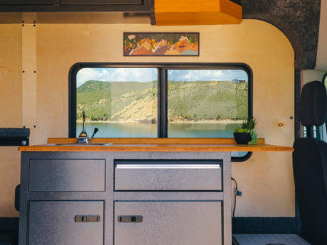 Inside an RV rental: a modern camper kitchen with sink, wooden countertop, and panoramic windows framing a serene lake and mountains.