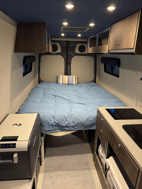 Compact rv rental interior: A cozy bed, mini kitchen with cooktop & sink, Dometic fridge, and overhead storage optimize this camper van.