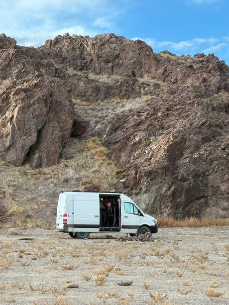 Adventure awaits! White camper van parked against rocky mountains, a person cheering from inside. Ideal rv rental for exploring nature.
