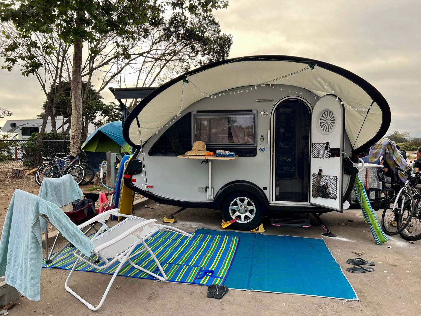 Relaxing at a campsite with a small RV rental (T@B travel trailer), featuring an awning, outdoor chairs & mats for comfort.