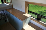 Bright RV rental interior featuring a bed, wood countertop, blue cabinets, and a window with a lush green view, ideal for travel.