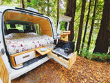 Inside a white camper van RV rental: a floral bed & pull-out kitchen with stove/sink, parked in a lush forest.