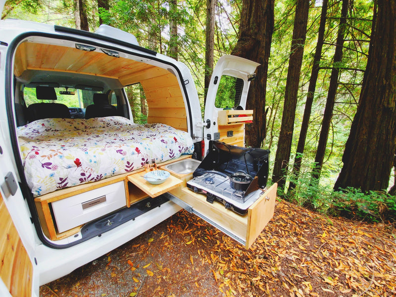 Inside a white camper van RV rental: a floral bed & pull-out kitchen with stove/sink, parked in a lush forest. — photo 7