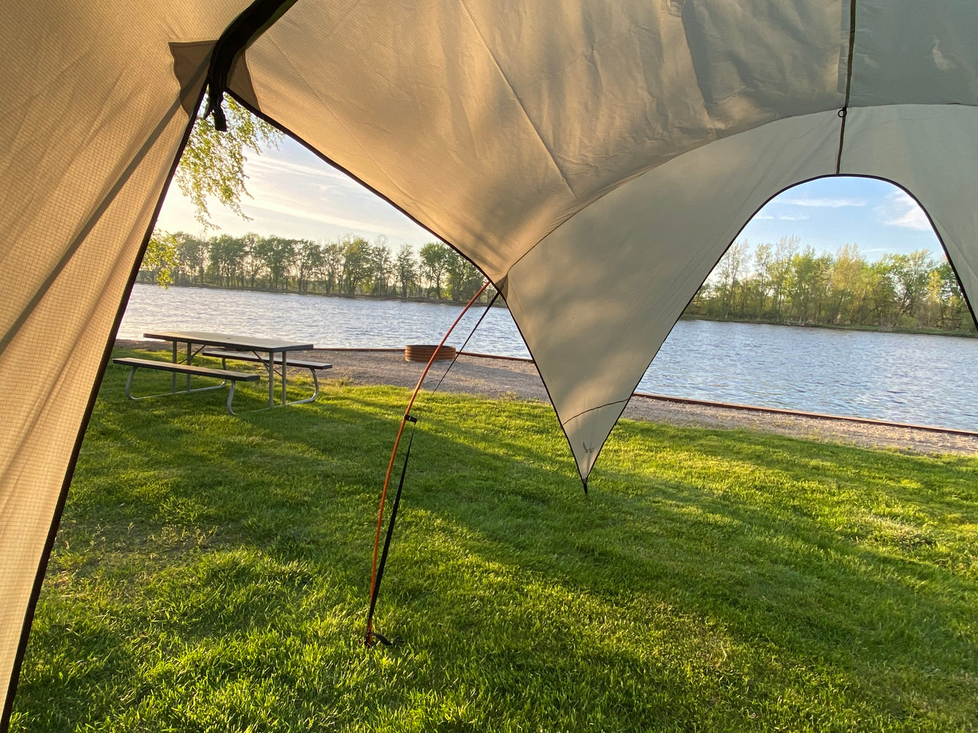View from an RV rental awning overlooking a serene lakeside campsite with a picnic table, fire pit, and lush grass. — photo 12