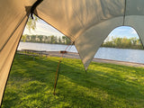 View from an RV rental awning overlooking a serene lakeside campsite with a picnic table, fire pit, and lush grass.