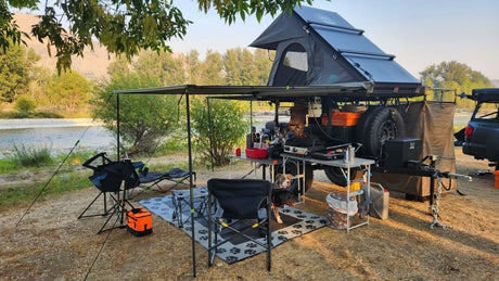 Fully equipped overlanding camp, like an RV rental, set by a scenic river. Features a rooftop tent, kitchen, awning, and a dog enjoying the outdoor adventure.