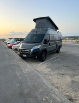 This grey Ram ProMaster rv rental, a pop-top camper van, is parked on sand beside a road, ready for adventure.