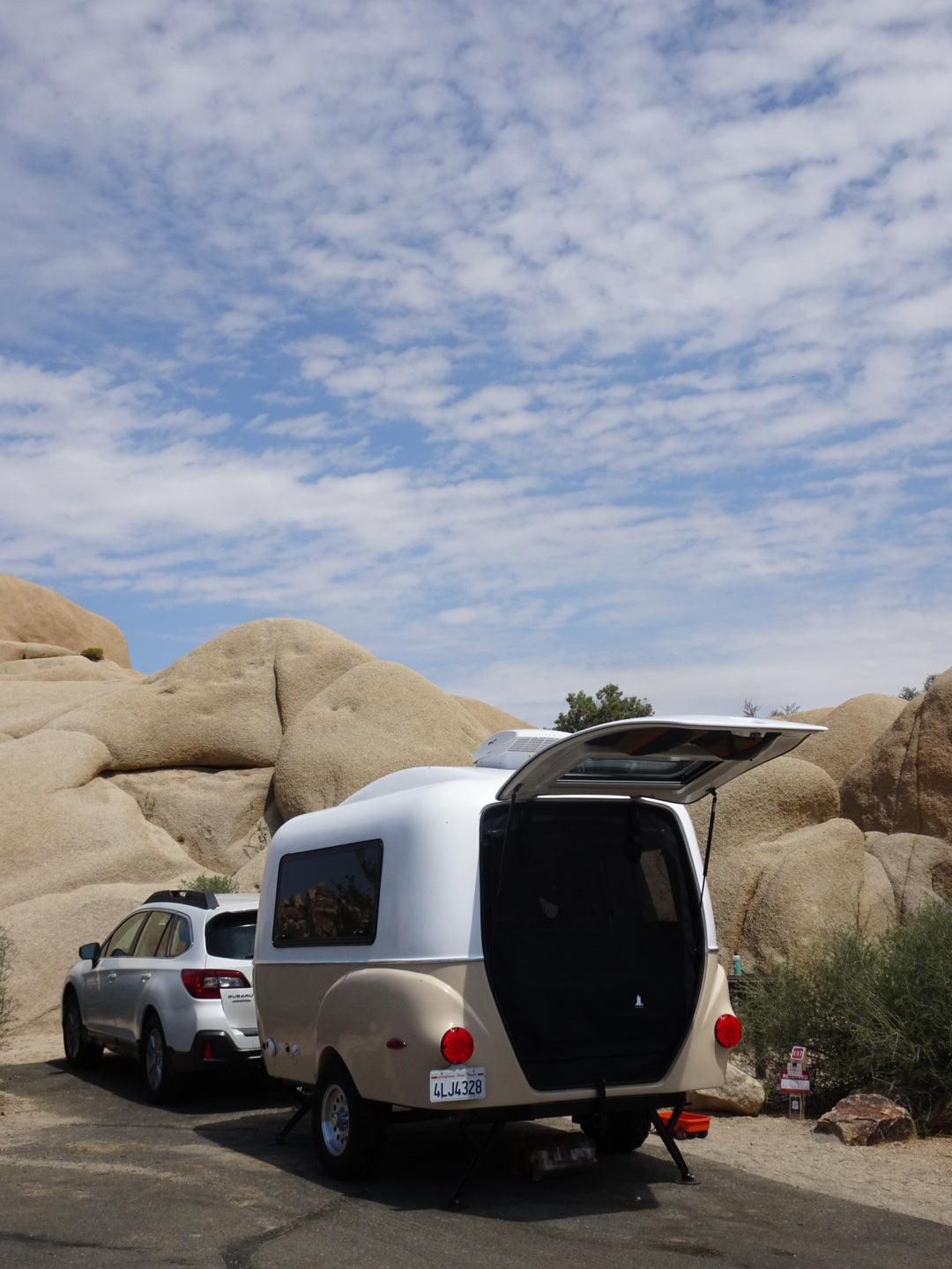 An RV rental, a small teardrop trailer, parked in a rocky desert, hatch open, blue sky above.