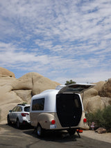 An RV rental, a small teardrop trailer, parked in a rocky desert, hatch open, blue sky above.