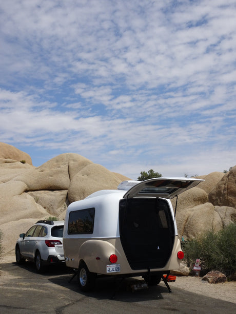 An RV rental, a small teardrop trailer, parked in a rocky desert, hatch open, blue sky above.