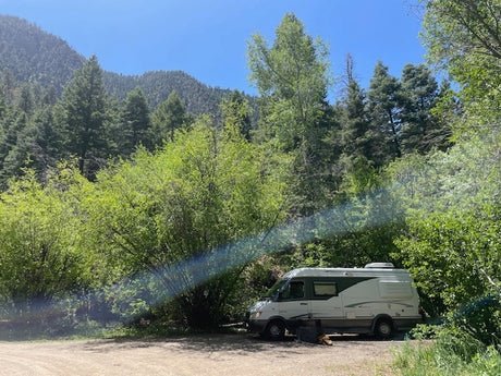 Experience nature with an RV rental! Camper van at a lush green forest campsite, surrounded by mountains under a blue sky.