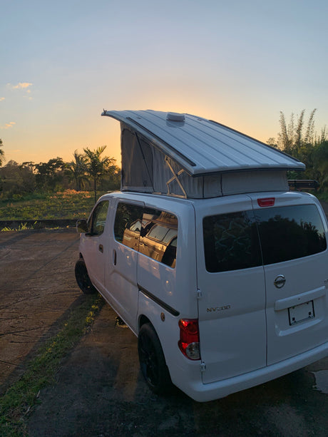 White Nissan NV200 camper van with pop-top extended at sunset, ready for an RV rental road trip in a tropical setting.