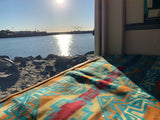 RV rental view: A patterned blanket frames bright sun reflecting on calm water, sandy shore, and distant palms.