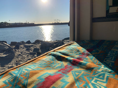 RV rental view: A patterned blanket frames bright sun reflecting on calm water, sandy shore, and distant palms.