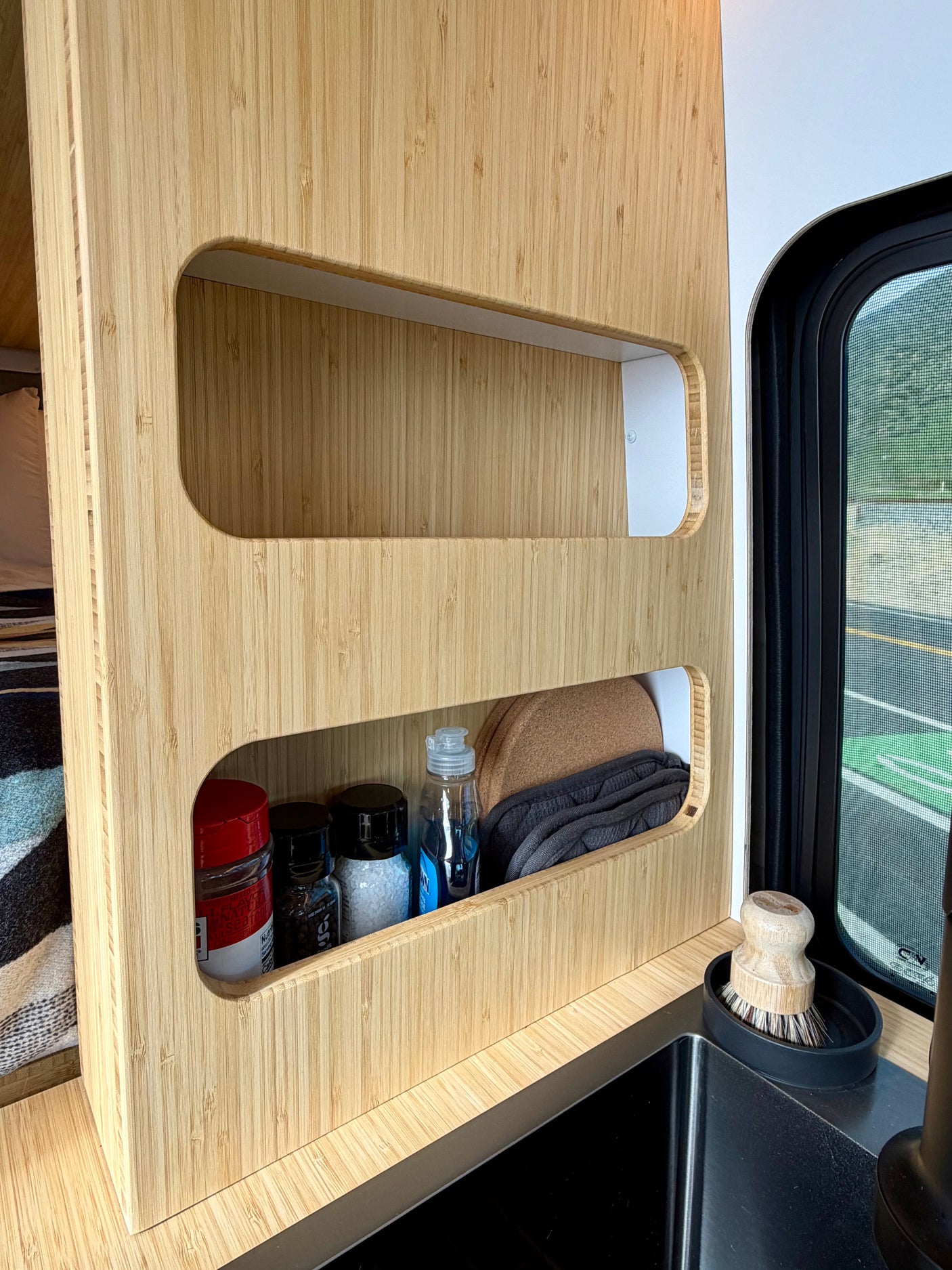 RV rental: Organized bamboo shelves in a van kitchen, holding spices, soap, and supplies by a sink and window.